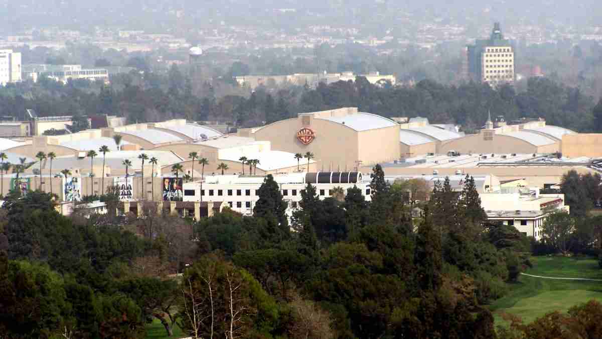 Aerial view of a large studio complex with a beige building bearing the Warner Bros shield, surrounded by trees and low structures.