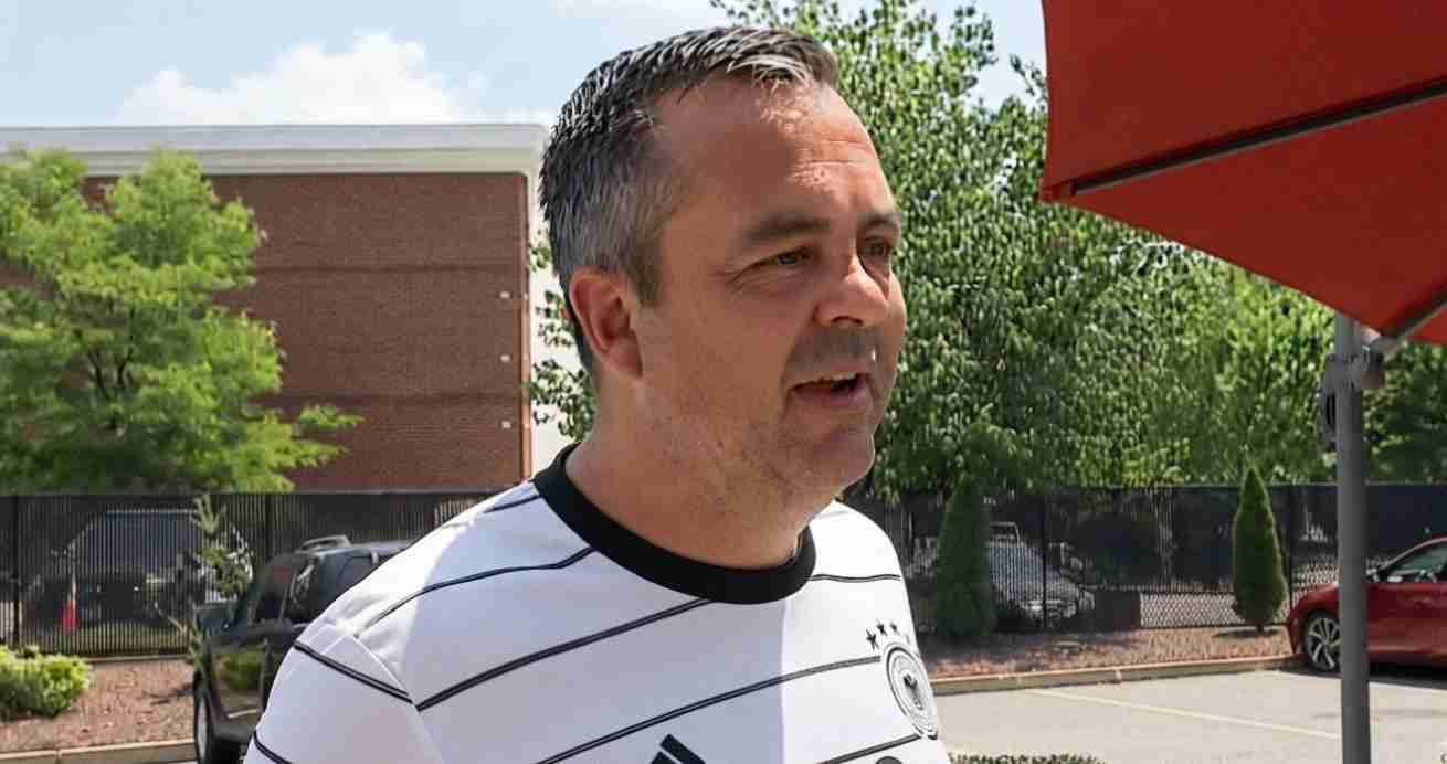 Middle-aged man in a white striped shirt speaking outdoors in a parking lot, with trees, a brick building, and a red umbrella in the background.