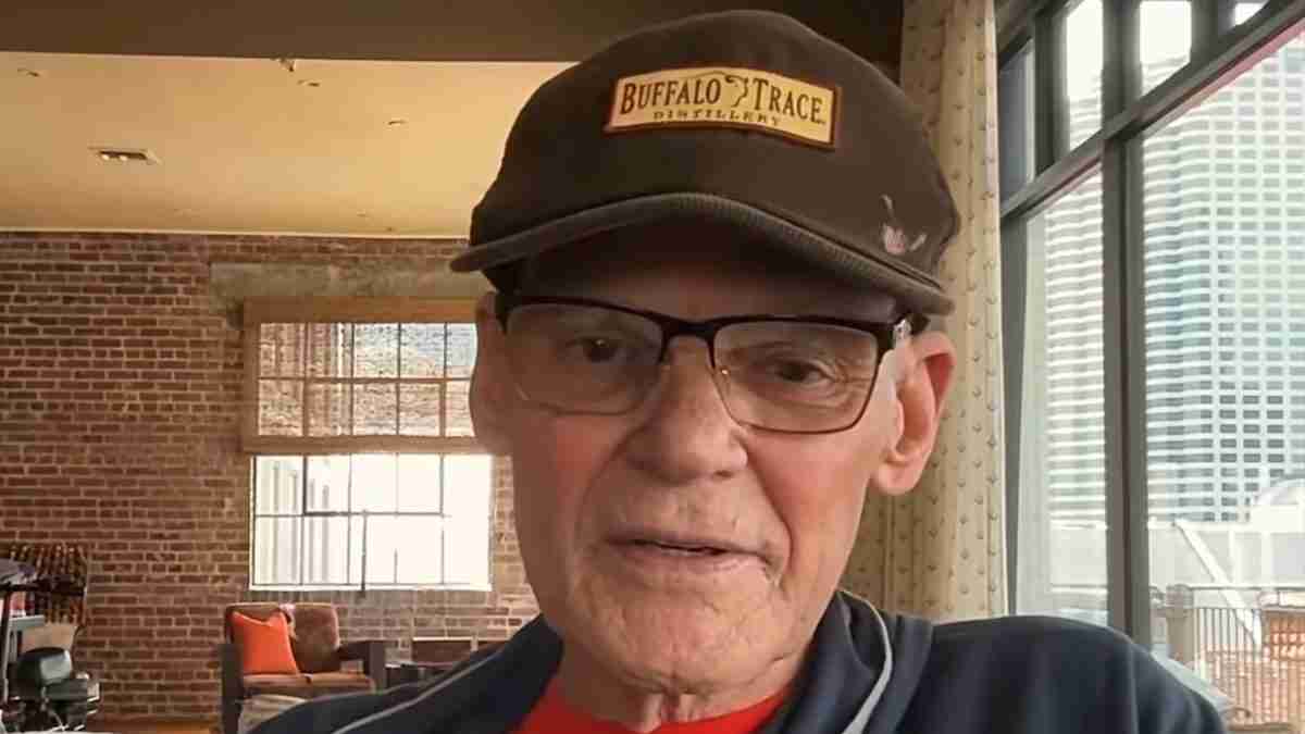 Close-up of an older man wearing glasses and a Buffalo Trace cap, indoors by a brick wall and large windows.