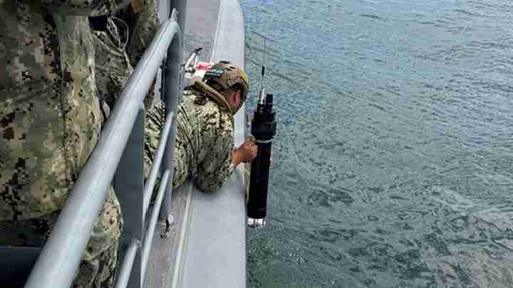 Military crew member in camouflage on a boat lowers a large black cylindrical device toward the water.