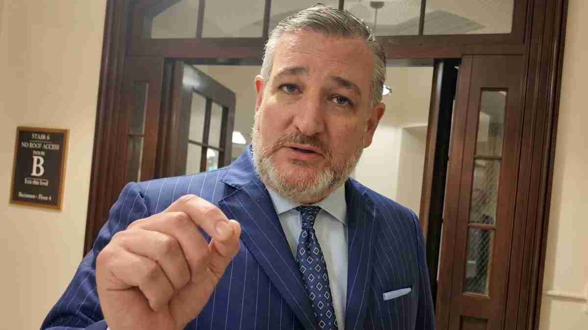 Man in a blue pinstripe suit raising his hand toward the camera in a hallway beside a wooden door.