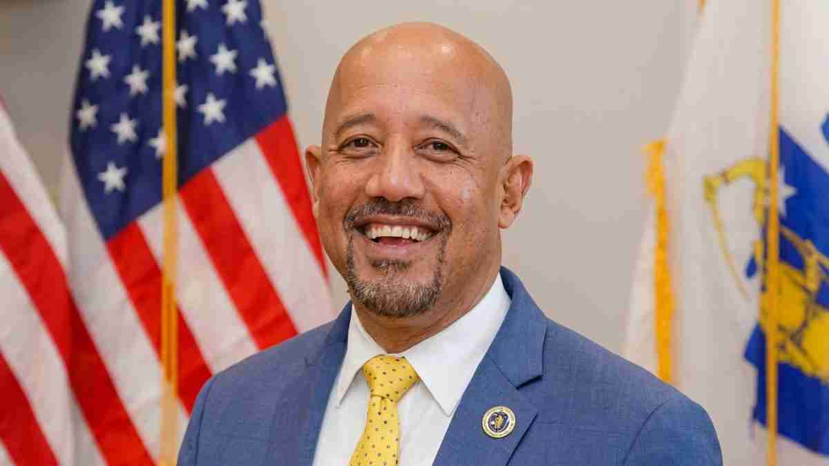 Smiling man in a blue suit and yellow tie, with American flags in the background behind him.