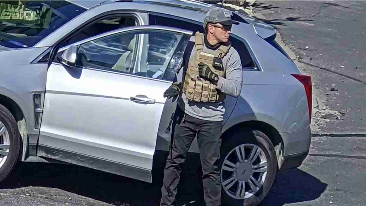Man in tactical vest and helmet stands next to a silver SUV with the driver door open.
