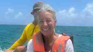 Smiling woman in an orange life jacket on a boat, with a man in a yellow jacket behind them on the blue ocean.
