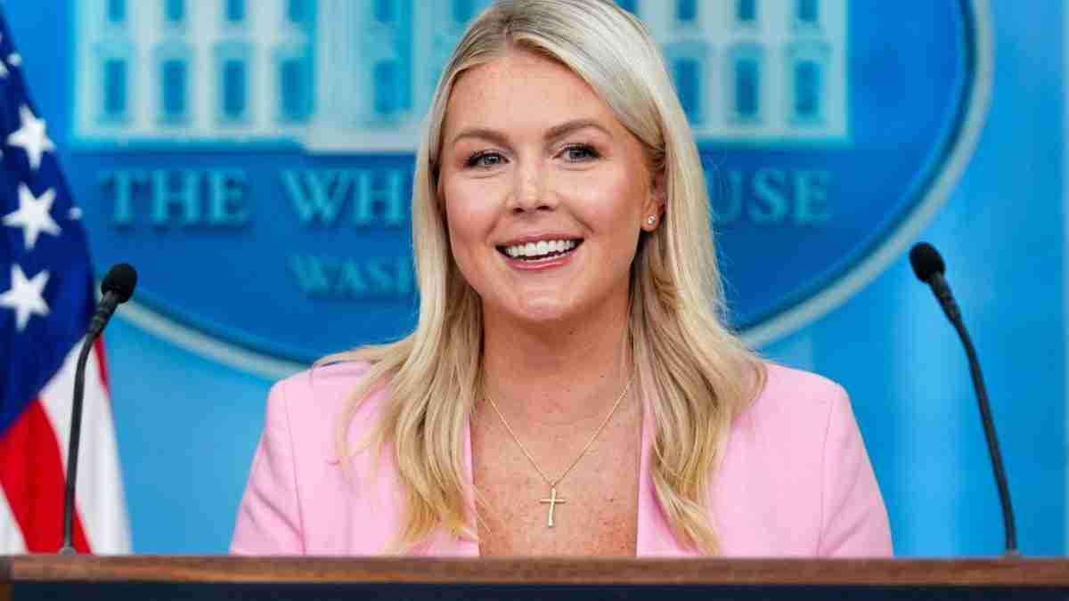 Woman at a press briefing behind a podium in the White House briefing room, wearing a pink blazer and a cross necklace, smiling at the camera.