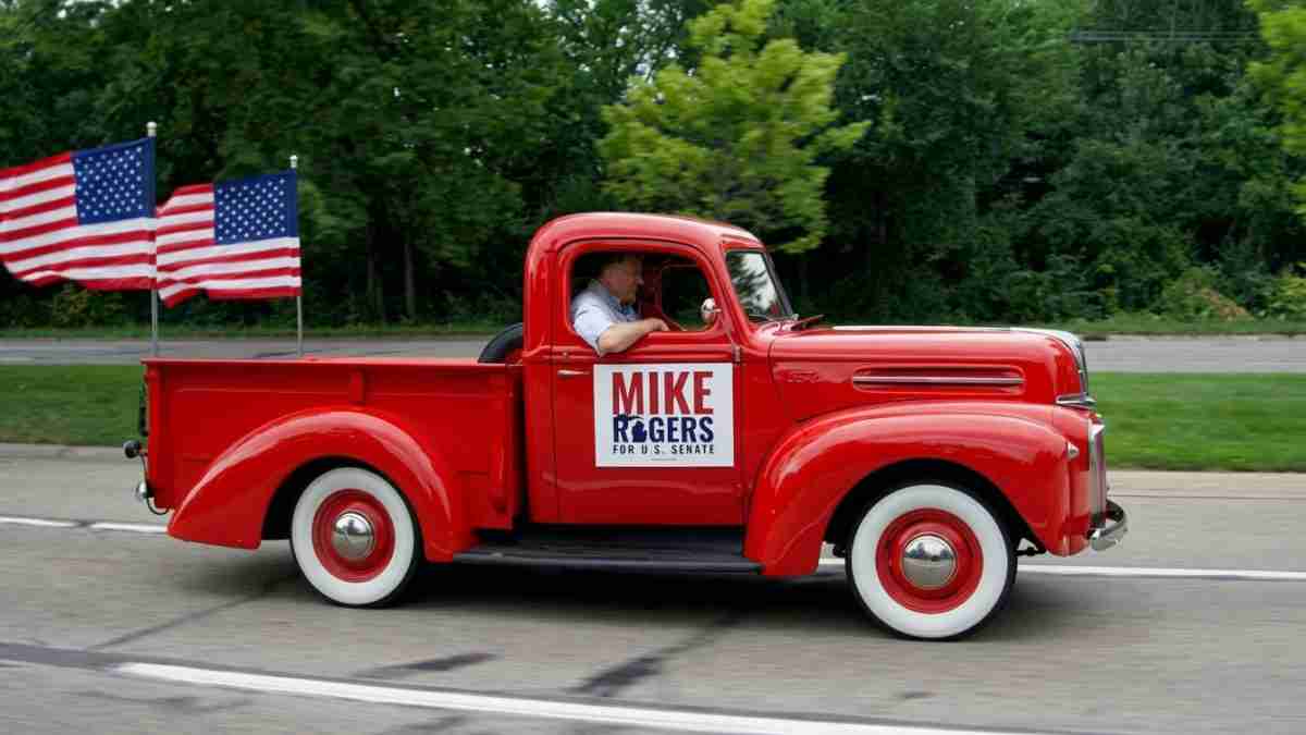 Vintage red pickup truck with a Mike Rogers for U.S. Senate campaign sign in the door, driver inside, two American flags in the bed behind him.