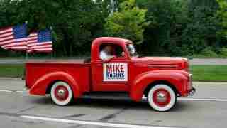 Vintage red pickup truck with a Mike Rogers for U.S. Senate campaign sign in the door, driver inside, two American flags in the bed behind him.