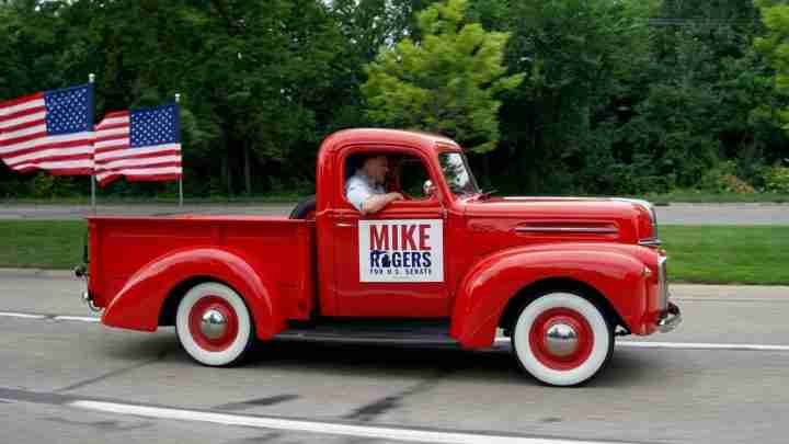 Vintage red pickup truck with a Mike Rogers for U.S. Senate campaign sign in the door, driver inside, two American flags in the bed behind him.