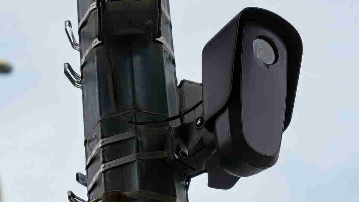 Outdoor CCTV camera mounted on a metal pole, angled to monitor the street against a cloudy sky.