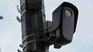 Outdoor CCTV camera mounted on a metal pole, angled to monitor the street against a cloudy sky.