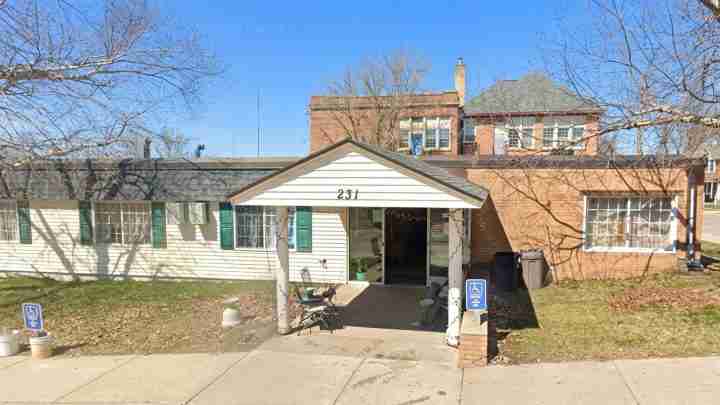 Single-story white building with a gabled front labeled 231, green shutters, a small porch with chairs and string lights, on a sunny day.