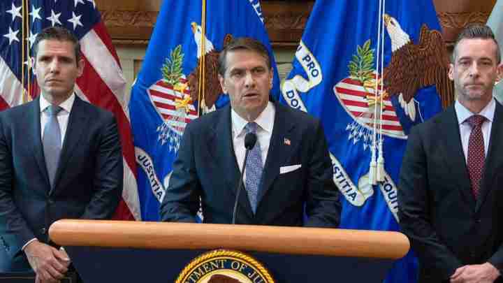 Three men in suits stand behind a podium with a Department of Justice seal, American and DOJ flags in the background.