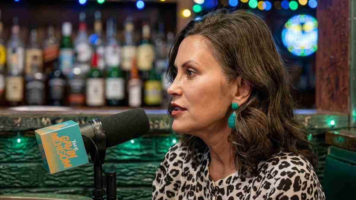 Woman in leopard-print top speaks into a microphone in a bar studio setting with colorful lights in the background.
