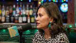 Woman in leopard-print top speaks into a microphone in a bar studio setting with colorful lights in the background.