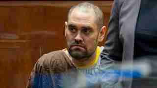 Man in a brown prison jumpsuit sits in a courtroom, looking to his right with a serious expression.