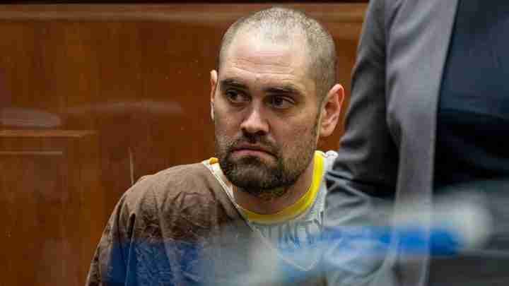 Man in a brown prison jumpsuit sits in a courtroom, looking to his right with a serious expression.