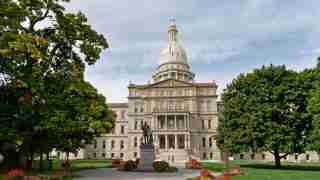 State capitol building with prominent dome, statue in front, and landscaped grounds with trees and flower beds