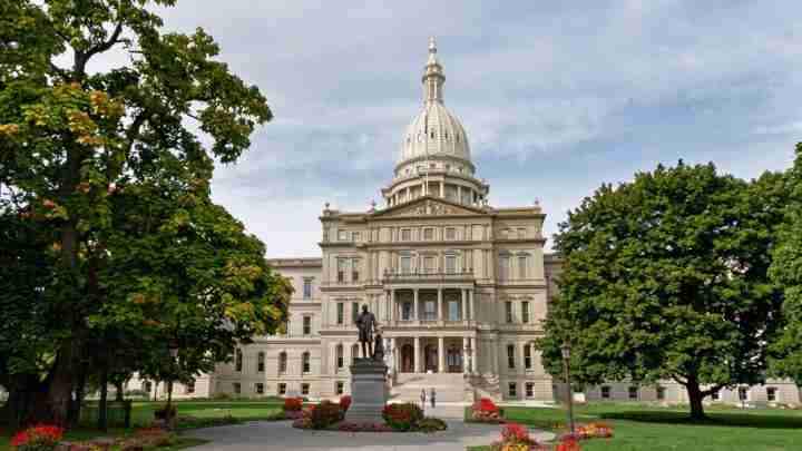 State capitol building with prominent dome, statue in front, and landscaped grounds with trees and flower beds
