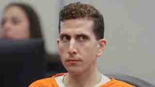 Man in an orange prison jumpsuit seated in a courtroom, looking to the side with a serious expression.