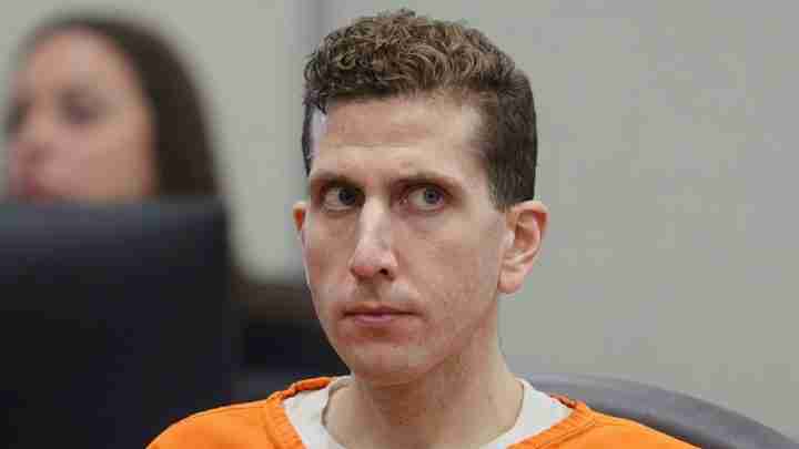 Man in an orange prison jumpsuit seated in a courtroom, looking to the side with a serious expression.