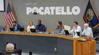 Six council members sit at a curved desk with laptops during a Pocatello city meeting; American and state flags in the background.