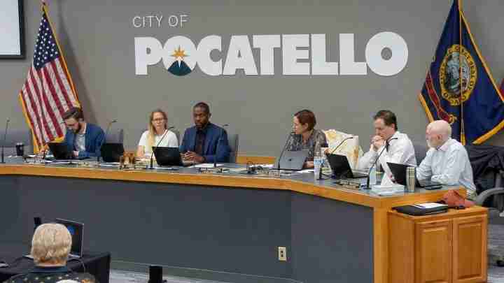 Six council members sit at a curved desk with laptops during a Pocatello city meeting; American and state flags in the background.