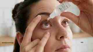 Close-up of a person administering eye drops: one hand steadies the eyelid while a drop falls toward the eye from a bottle above it.