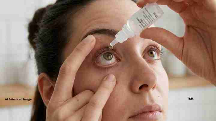 Close-up of a person administering eye drops: one hand steadies the eyelid while a drop falls toward the eye from a bottle above it.