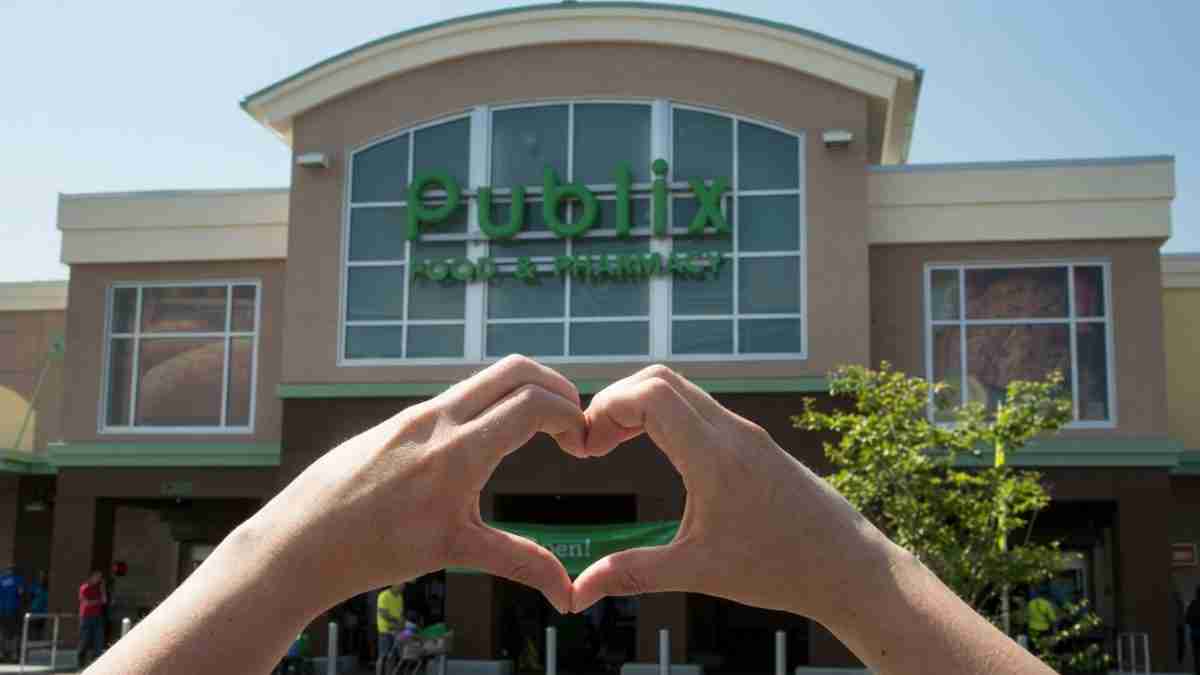 Two hands forming a heart shape in front of a large storefront with glass windows on a sunny day.