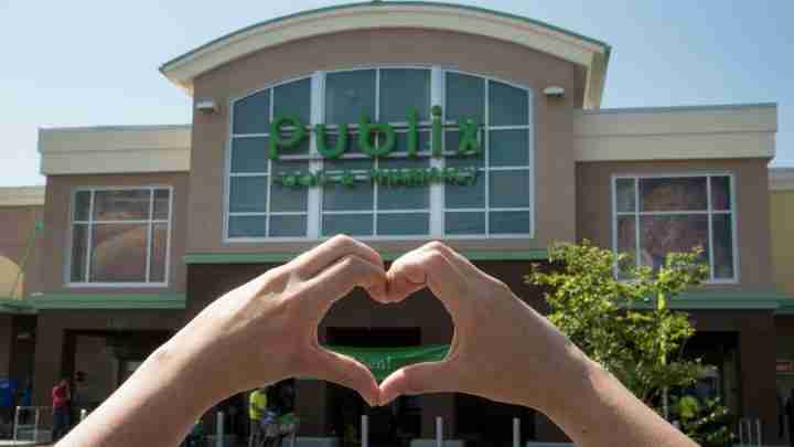 Two hands forming a heart shape in front of a large storefront with glass windows on a sunny day.