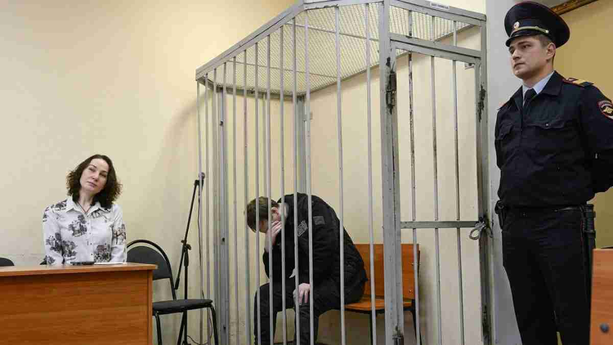 A woman sits at a courtroom desk while a man in a jail cell hides his face, with a uniformed guard standing nearby.