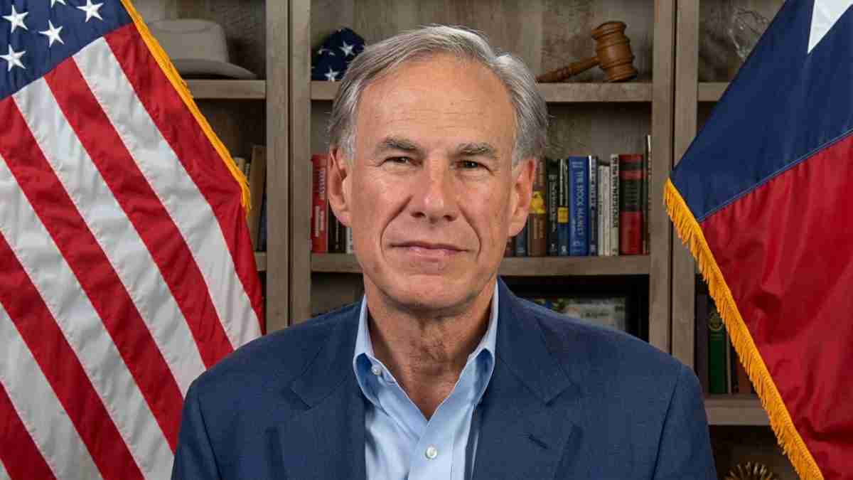 Man in a blue blazer and light blue shirt facing the camera, standing in an office with American and state flags visible behind him.