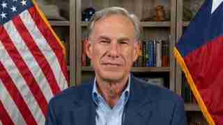 Man in a blue blazer and light blue shirt facing the camera, standing in an office with American and state flags visible behind him.