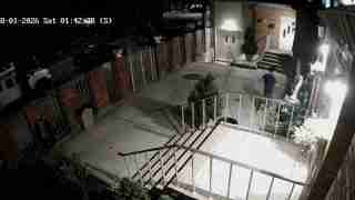 Night exterior of a brick building with a person in a blue shirt walking toward the entrance; stairs and railing lead to a courtyard; timestamp 08-01-2026 Sat 01:42:38 (S).