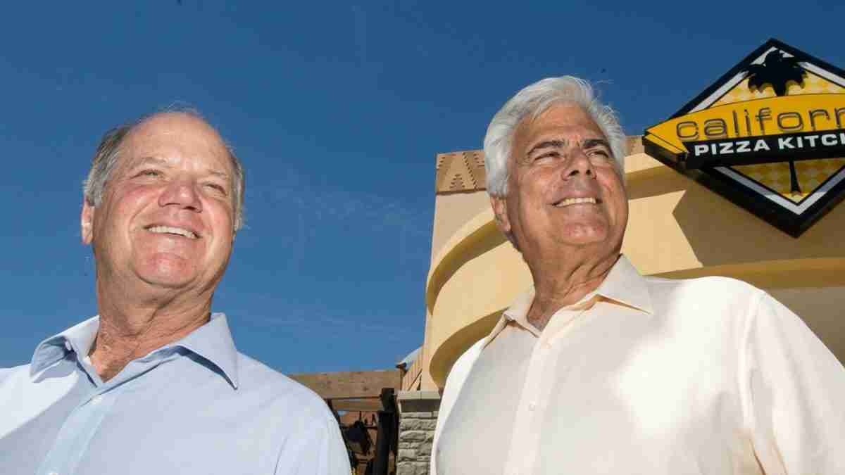 Two smiling older men in light shirts stand outdoors near a California Pizza Kitchen sign in the background.