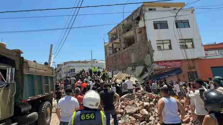 Street scene after a building collapse with rubble, a crowd of onlookers, and rescue workers in helmets coordinating work by a collapsed façade behind them.