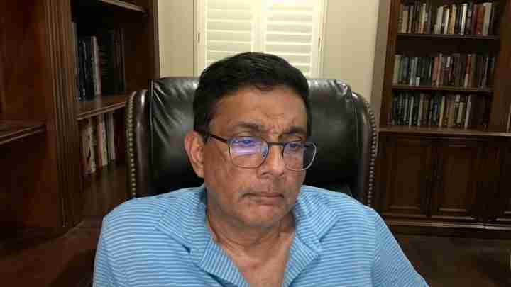 Man wearing glasses sits in a leather chair in a home library with dark wood bookshelves behind him.