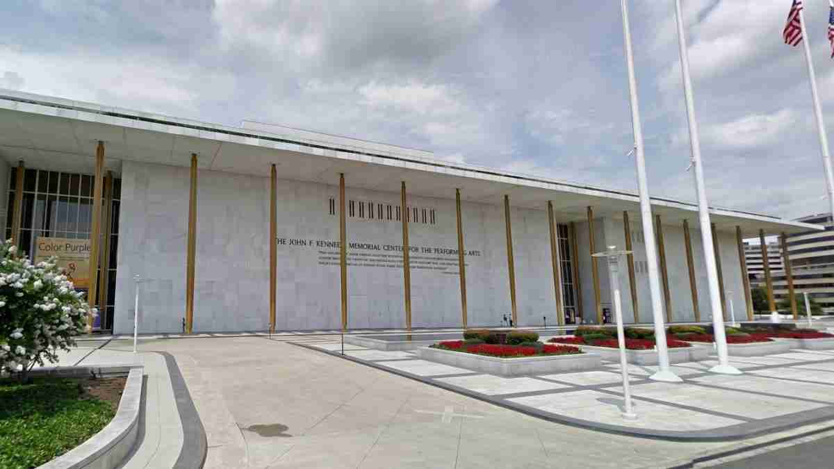 Modern public building with tall gold columns and a wide entrance, flagpoles on the right, and manicured gardens in front, likely a memorial center.