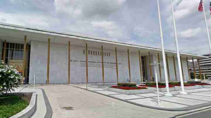 Modern public building with tall gold columns and a wide entrance, flagpoles on the right, and manicured gardens in front, likely a memorial center.