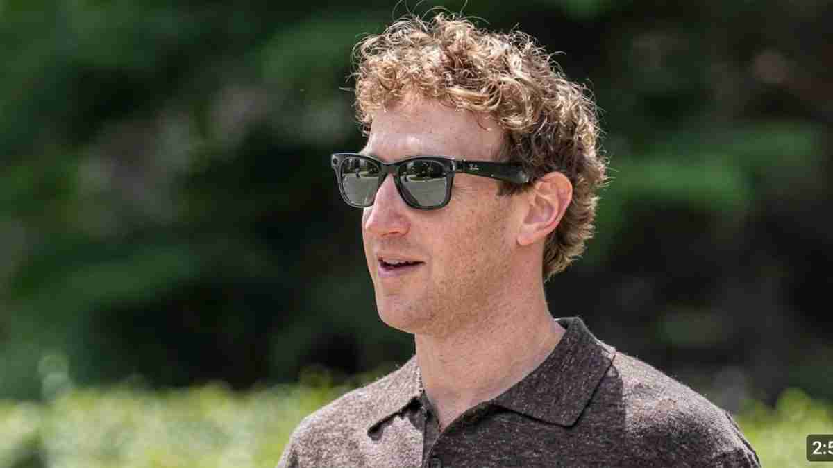 Man with curly hair wearing dark sunglasses, looking to the right, outdoors with greenery in the background.