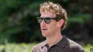 Man with curly hair wearing dark sunglasses, looking to the right, outdoors with greenery in the background.